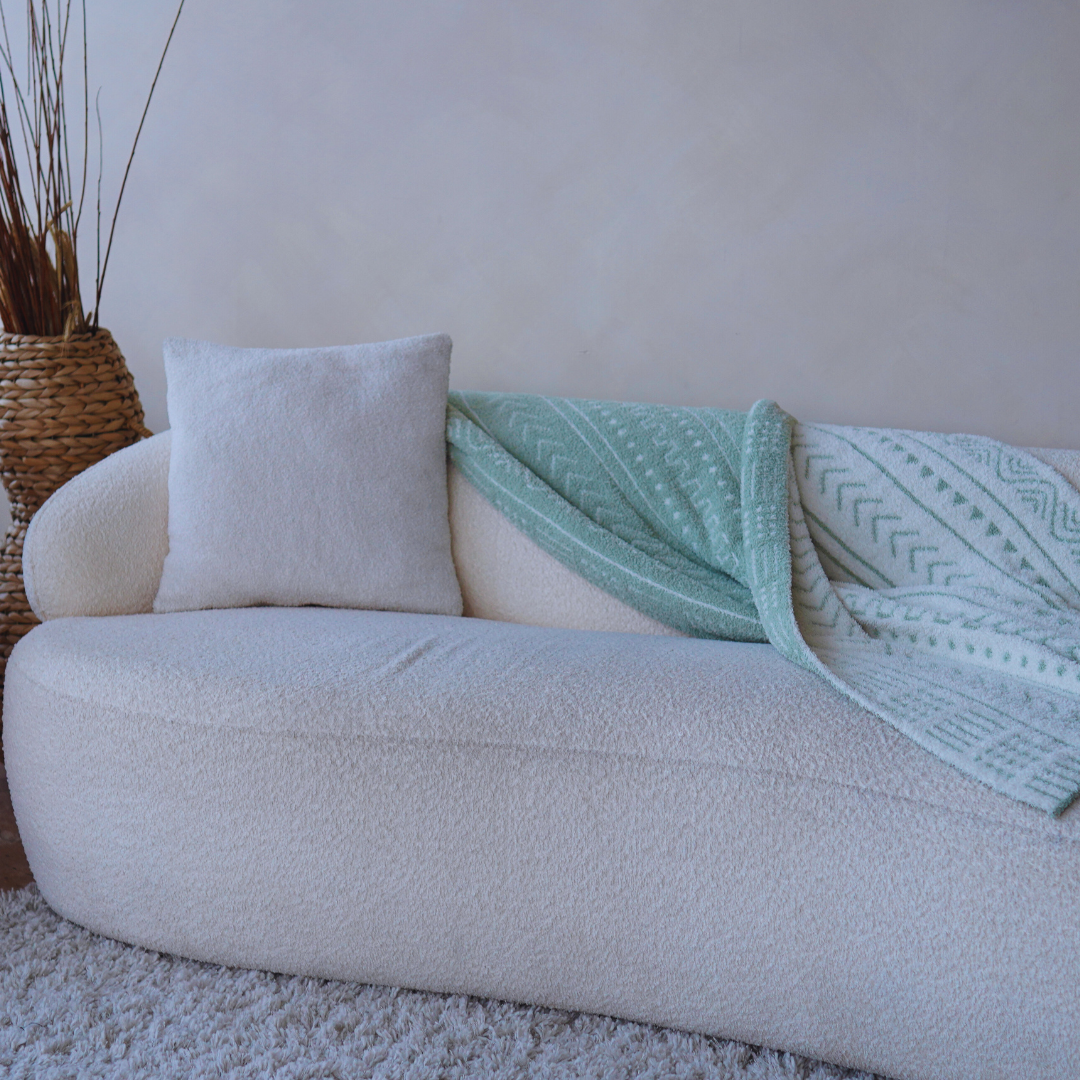 Minimalist cream sofa with a textured throw pillow and a folded mint green patterned blanket draped across, placed against a neutral wall with a woven floor vase nearby