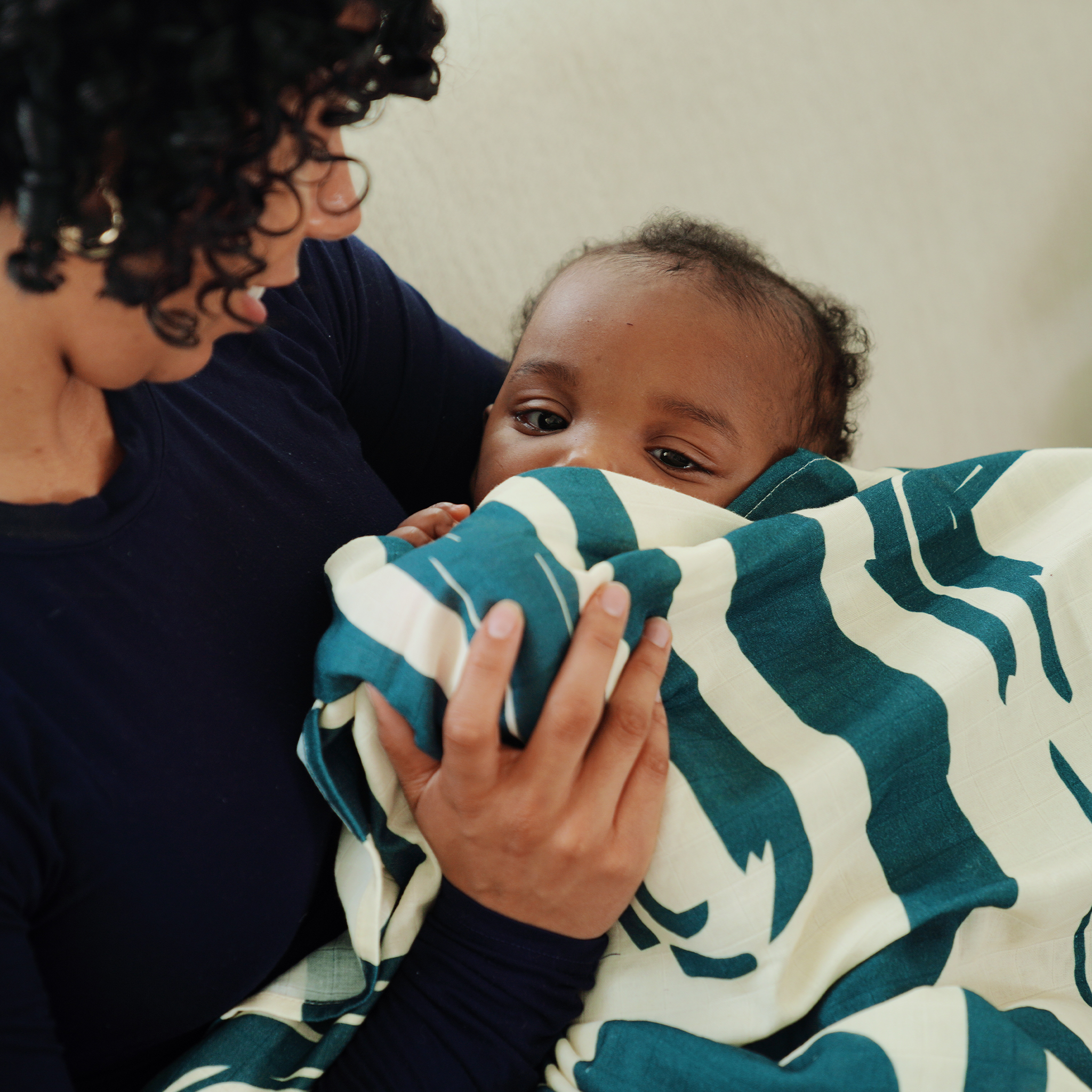 Mother holding her baby wrapped in a cream and teal abstract patterned blanket, sharing a quiet bonding moment on a sofa