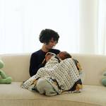 Mother bottle-feeding her baby while sitting on a beige sofa, wrapped in a patterned blanket in a bright, cozy living room