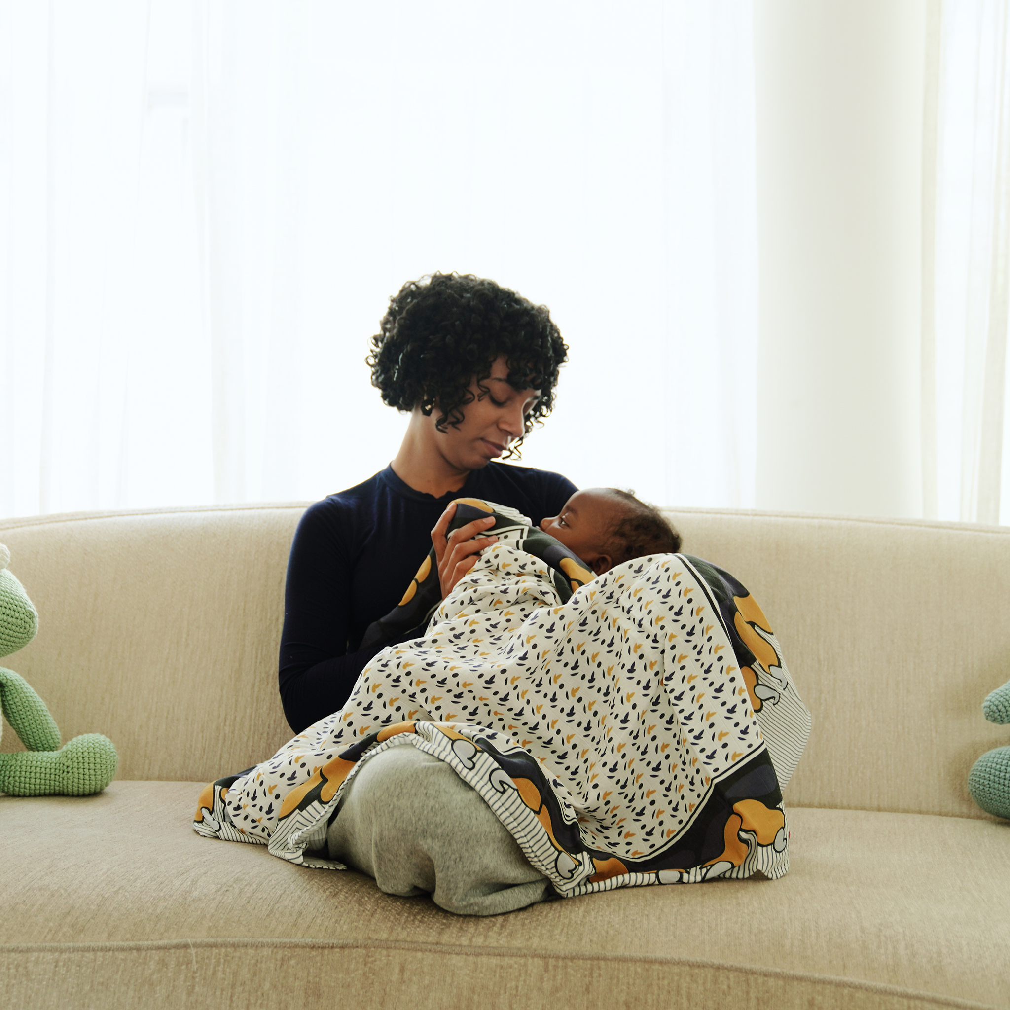 Mother bottle-feeding her baby while sitting on a beige sofa, wrapped in a patterned blanket in a bright, cozy living room