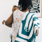 Mother carrying her baby with a bold, abstract-patterned green and cream blanket draped over her shoulder, standing in a modern home with framed art on the wall