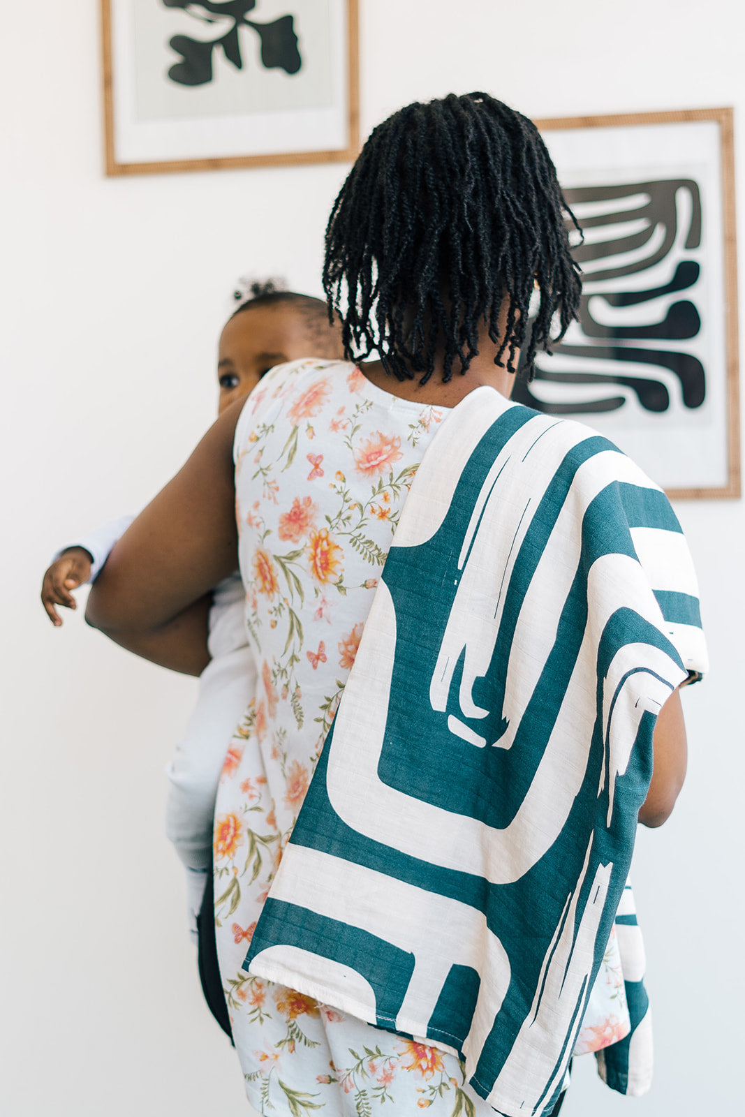 Mother carrying her baby with a bold, abstract-patterned green and cream blanket draped over her shoulder, standing in a modern home with framed art on the wall