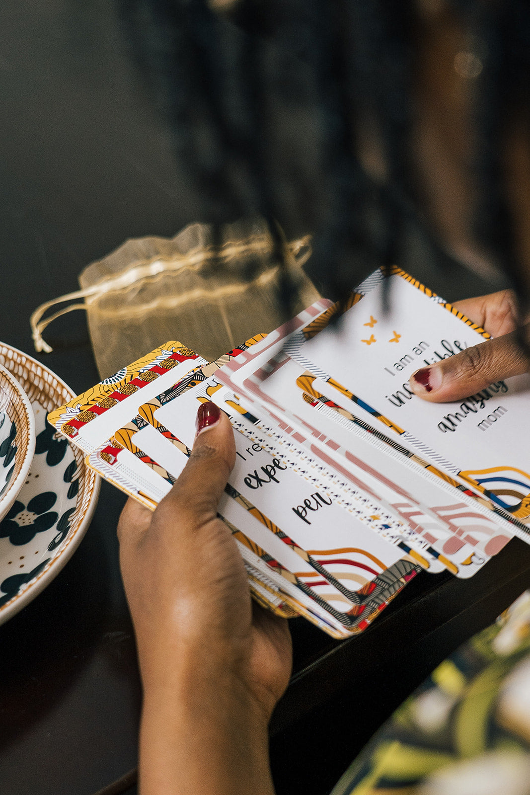 Close-up of a woman’s hands flipping through a colorful deck of affirmation cards with empowering messages for moms, next to a drawstring pouch on a table