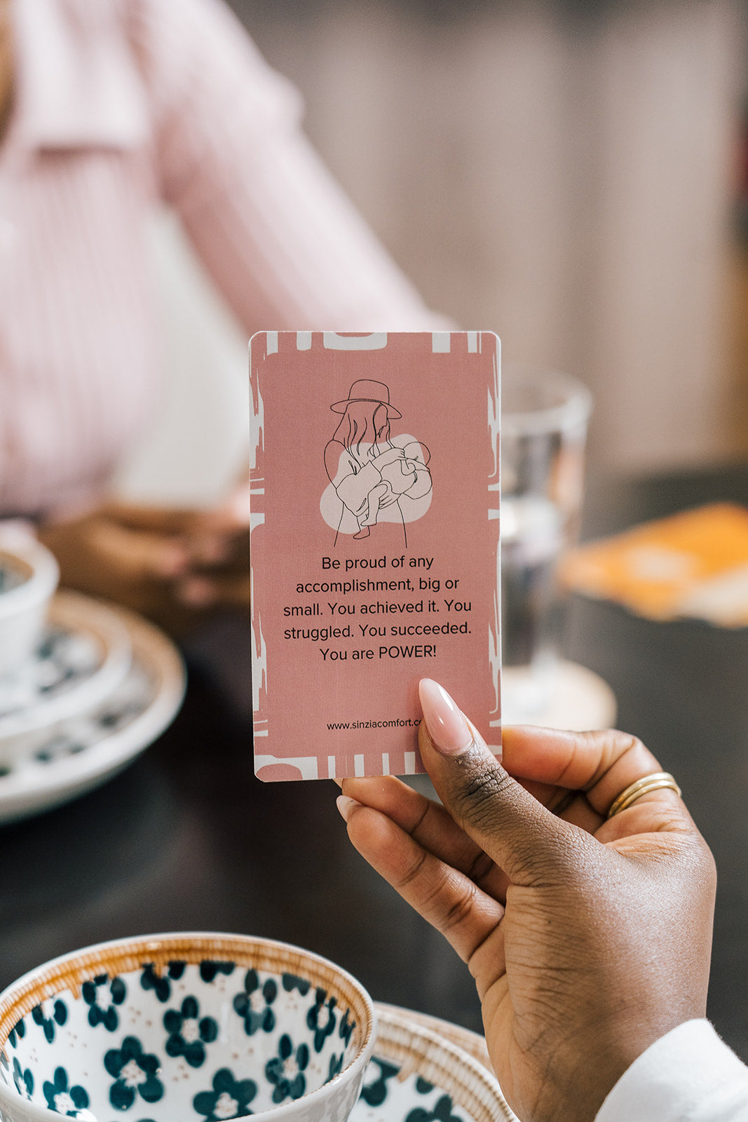 Close-up of a hand holding an affirmation card that reads 'Be proud of any accomplishment...' during a moment of encouragement at the dining table
