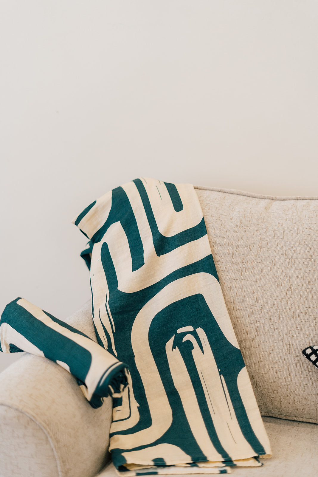 Beige armchair with a folded abstract cream and dark green patterned throw blanket draped over the side, in a minimal living space