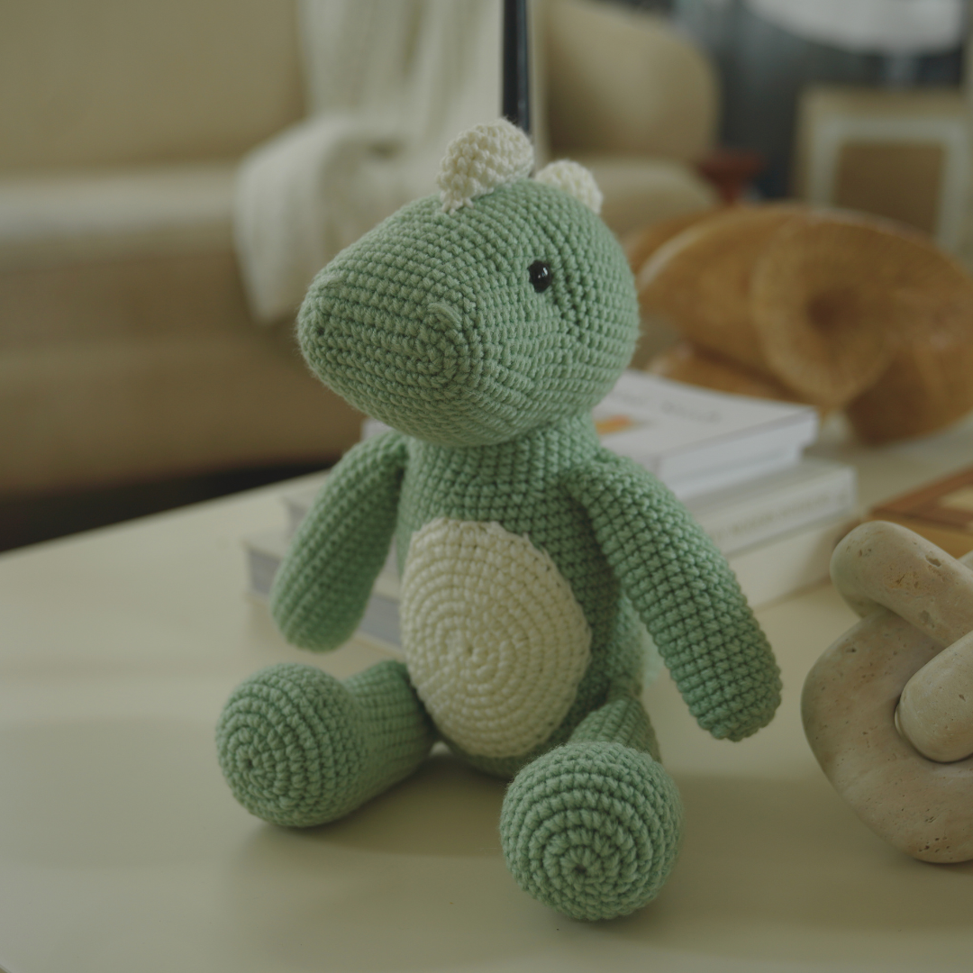 Handmade green crochet dinosaur toy sitting on a coffee table in a cozy living room with books and decorative objects