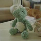 Handmade green crochet dinosaur toy sitting on a coffee table in a cozy living room with books and decorative objects