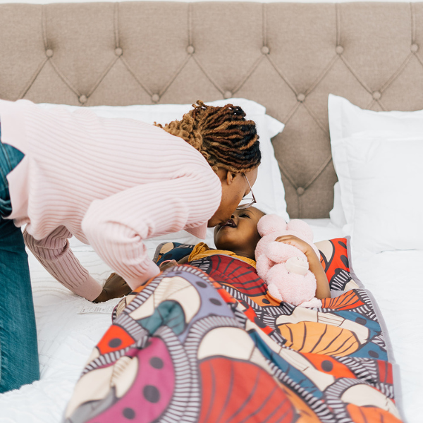 Mother leaning in to kiss her daughter goodnight as she lies in bed with a colorful patterned blanket and stuffed animal