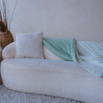 Minimalist cream sofa with a textured throw pillow and a folded mint green patterned blanket draped across, placed against a neutral wall with a woven floor vase nearby