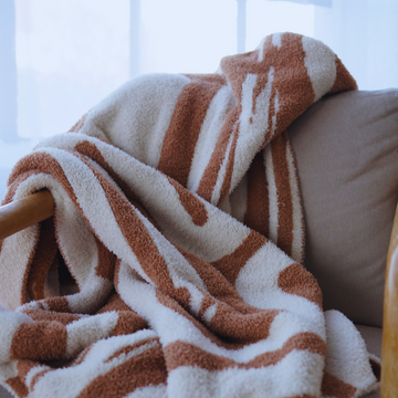 Soft terracotta and cream abstract-patterned blanket draped over a wooden armchair in a cozy, sunlit living room