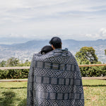 Two people wrapped in a patterned blanket overlooking a scenic landscape with mountains and trees.