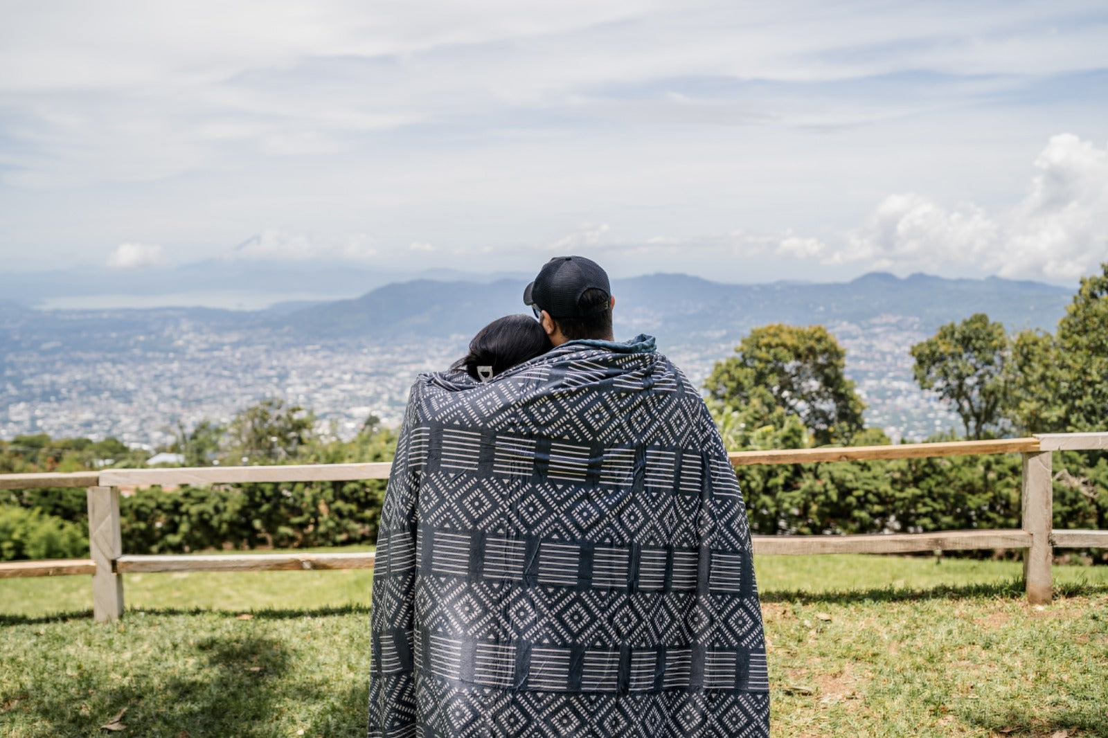 Two people wrapped in a patterned blanket overlooking a scenic landscape with mountains and trees.