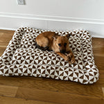 Small brown dog lying on a geometric-patterned pet bed on hardwood floor next to a white wall